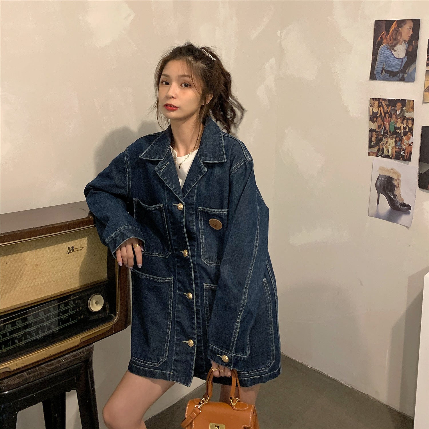 Woman wearing a denim jacket standing next to a vintage radio in a room with posters on the wall.
