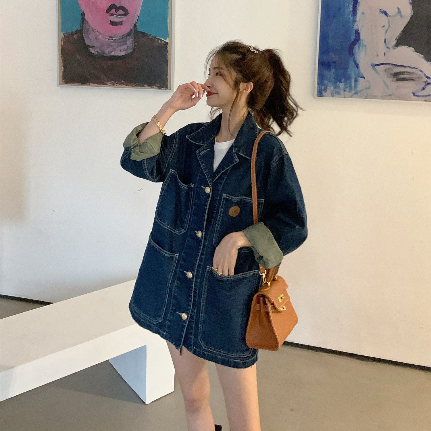 Woman wearing a denim jacket and holding a brown handbag in an art gallery.