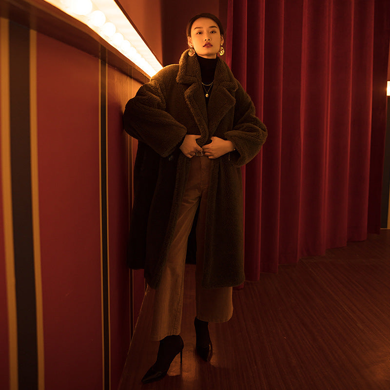 Person wearing a long brown coat in a dimly lit room with red curtains.